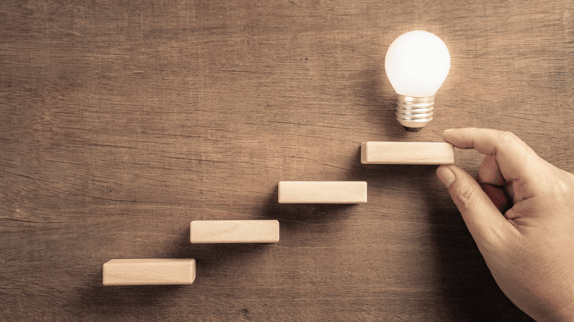 A hand placing wooden blocks as steps toward a glowing light bulb – illustrating how Google Ads work in a step-by-step approach.